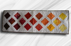 Handmade quilted autumn table runner with orange, red, gold, green, and brown diamond patchwork on a white quilted background.