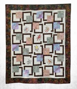 Handmade embroidered bird quilt with pastel batik patchwork blocks, cream quilting, dark brown geometric sashing, and a wide woodland floral border.