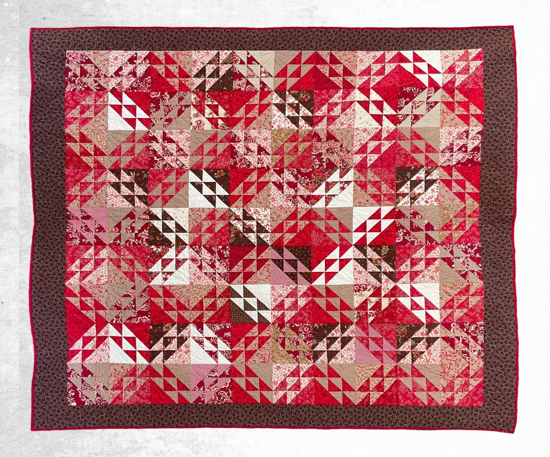 Handmade king patchwork quilt in red, brown, tan, cream, and white triangle blocks with a dark brown floral border and red binding, shown flat from above.