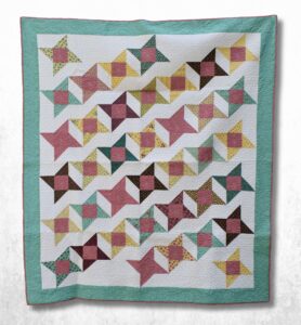 A flat lay view of a square-patterned cotton quilt featuring multicolored geometric stars on a white background, enclosed by a solid mint green outer border.