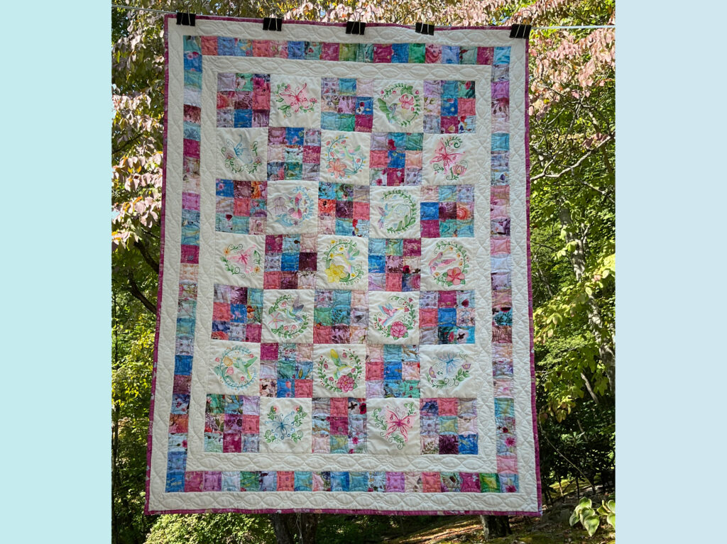 Quilt featuring beautifully embroidered butterfly and hummingbird motifs, framed by a patchwork of floral patchwork squares in pinks, blues, and purples,