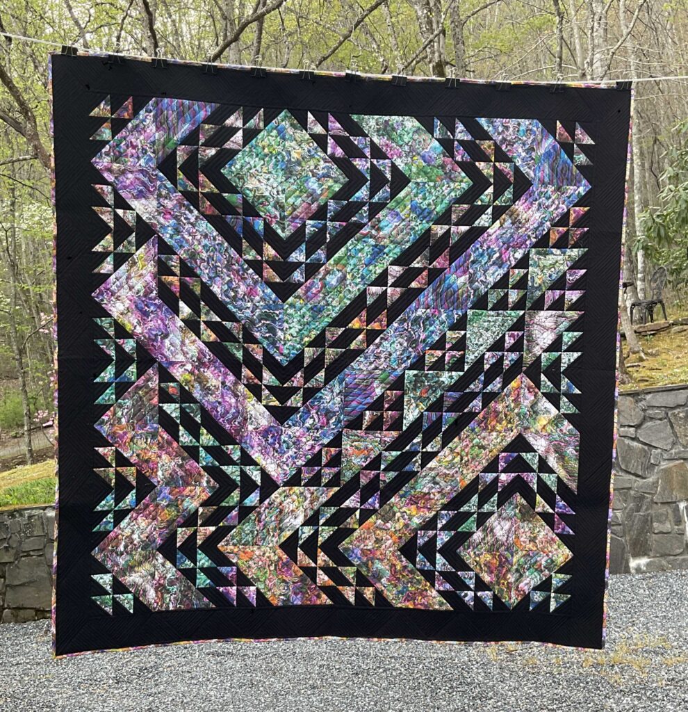 Bold, abstract handmade quilt features vivid fabrics in swirling jewel tones, set against a deep black background.