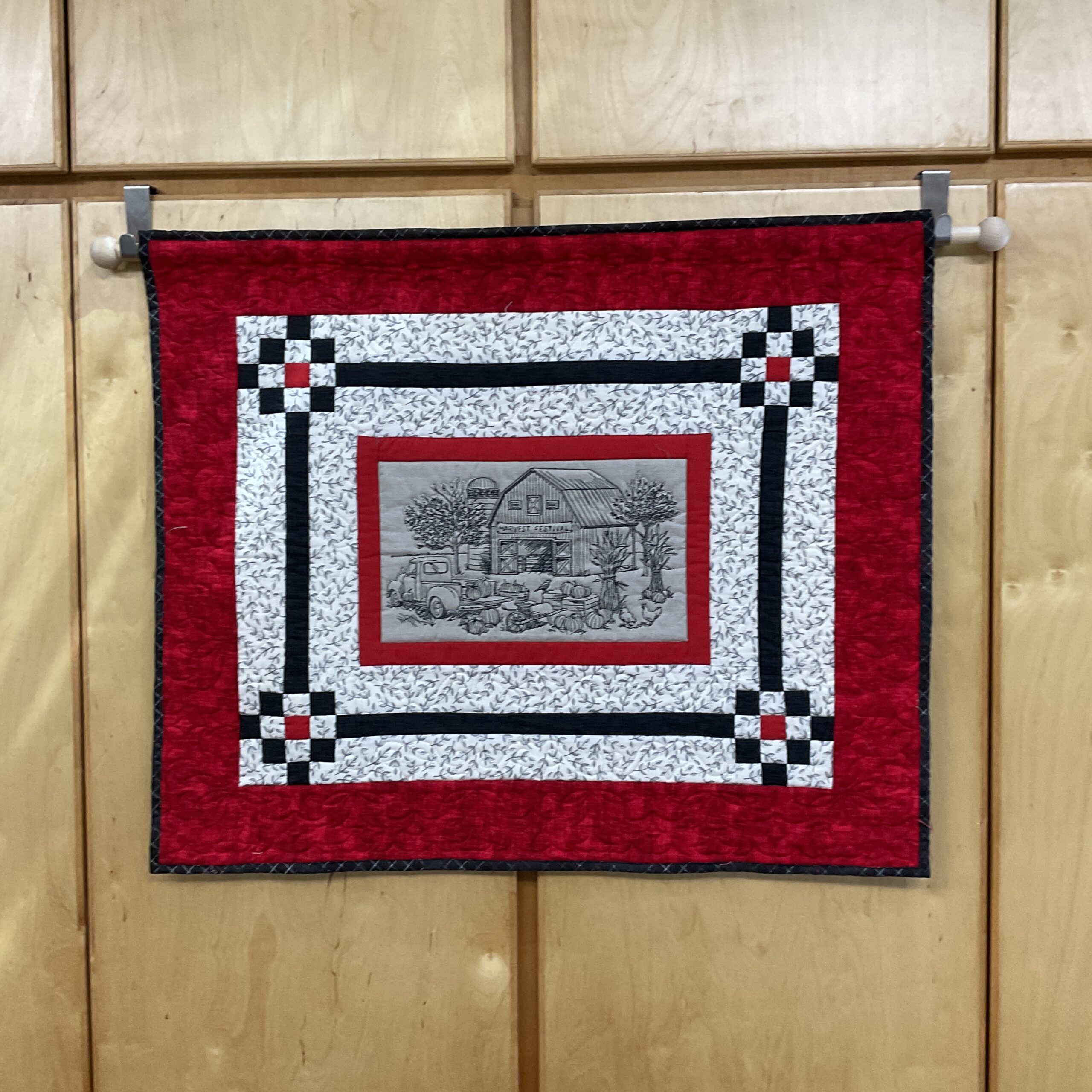 Horizontal 32 x 27 inch quilted wall hanging with an embroidered harvest festival farm scene, red outer border, cream floral background, black geometric framing, and checkerboard corner accents.