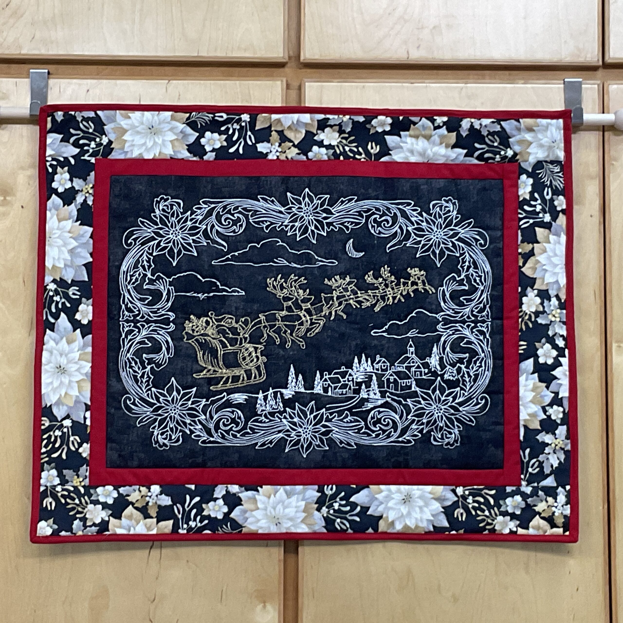 Horizontal 32 x 26 inch quilted Christmas wall hanging with embroidered Santa and reindeer flying over a winter village, metallic gold accents, red framing, and a black poinsettia floral border.