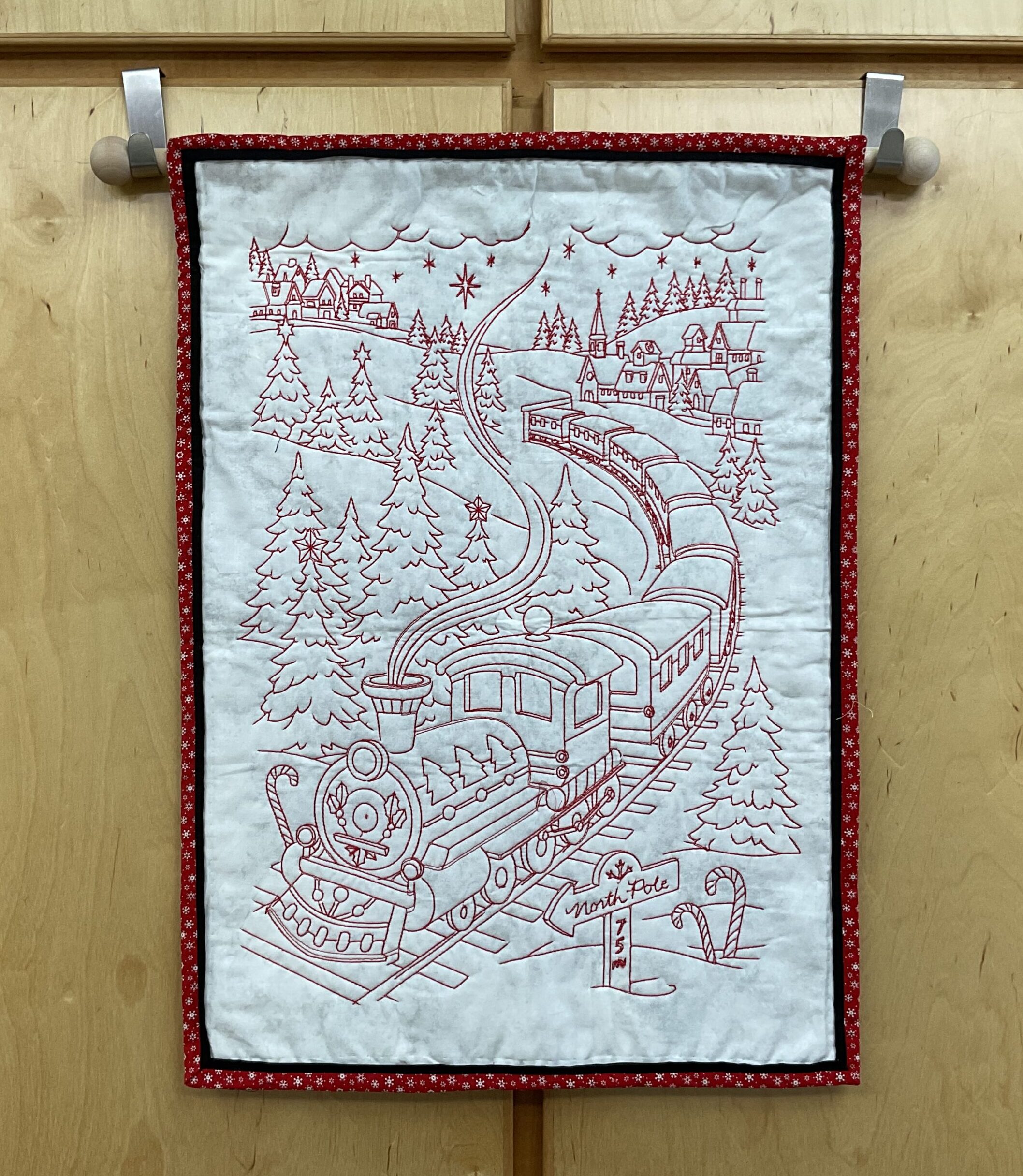 Vertical 18 x 26 inch quilted Christmas wall hanging with red embroidered steam train, snowy village, evergreen trees, candy canes, and North Pole sign on a pale background.