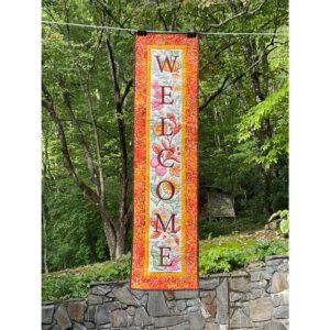 Tall 16 x 61 inch quilted wall hanging with embroidered letters spelling WELCOME, decorated with autumn leaves, flowers, and pumpkins, framed by gold and bright orange borders.