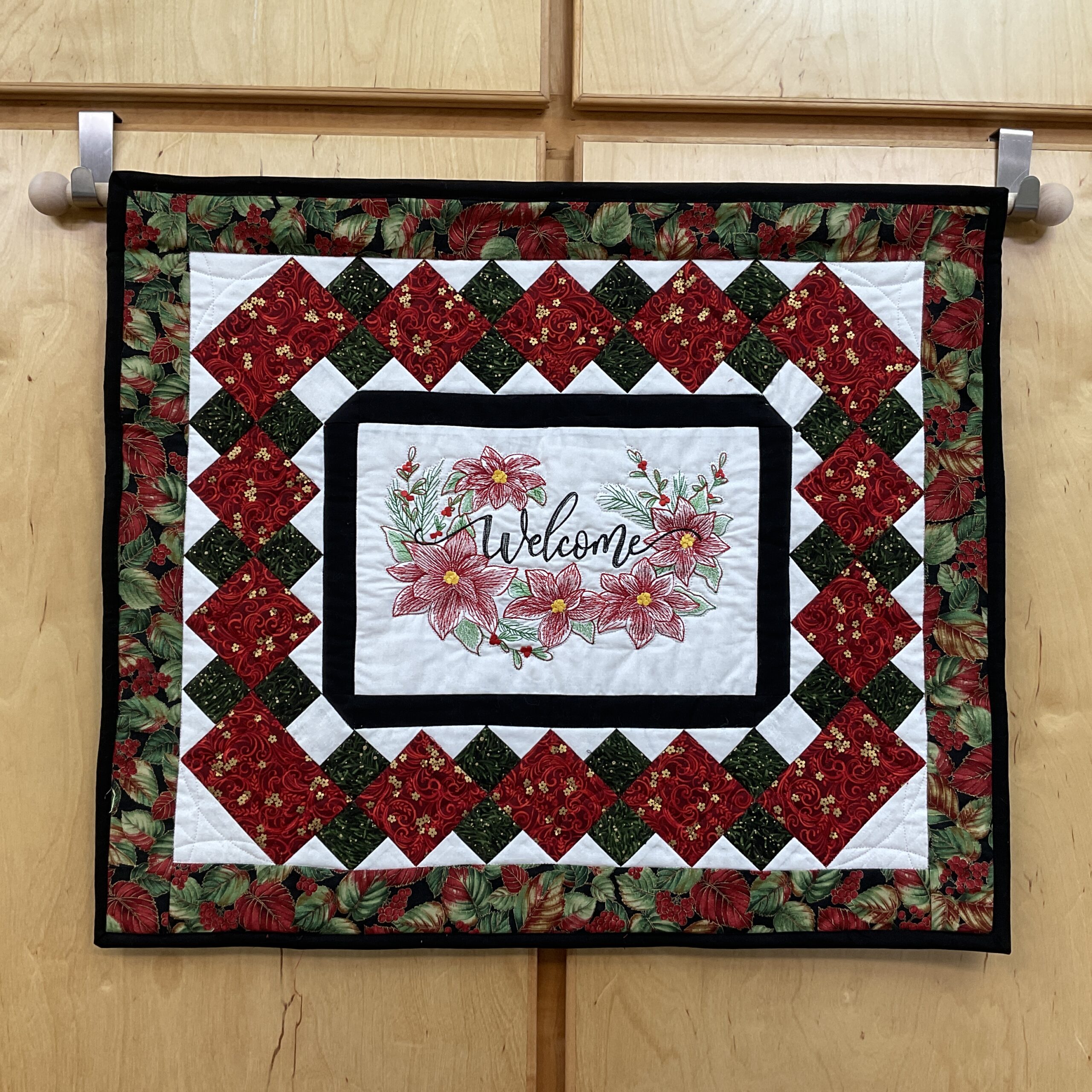 Horizontal 26 x 22 inch quilted Christmas wall hanging with embroidered poinsettias and “Welcome†lettering, framed by red and green diamond patchwork, black accents, and a dark floral poinsettia border.