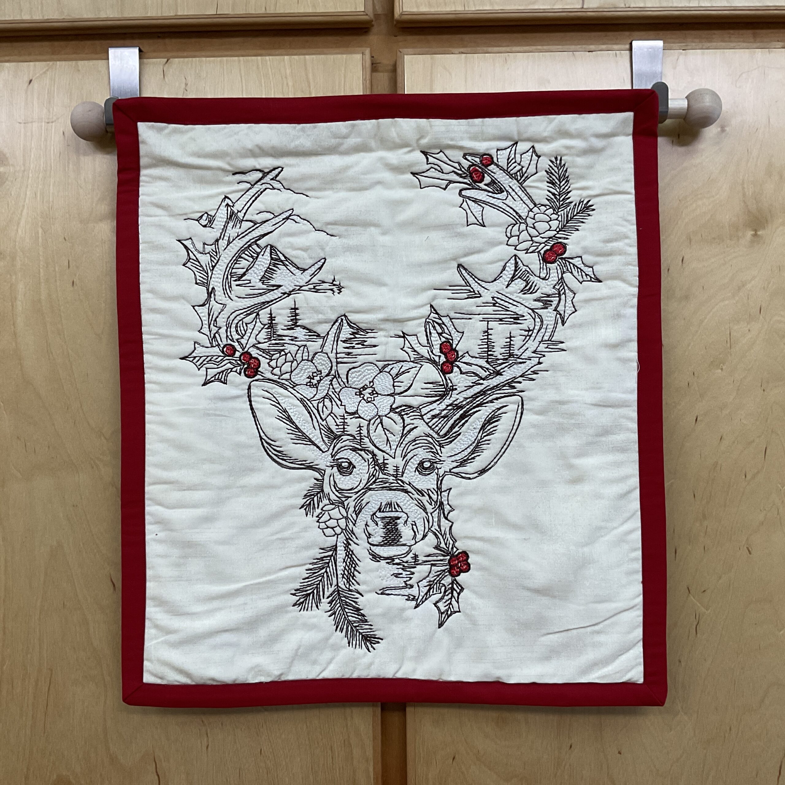 Vertical 17 x 20 inch quilted wall hanging with an embroidered stag, winter branches, holly, pinecones, red berries, and red binding on a cream background.