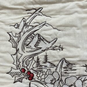Close-up of Christmas Stag embroidery showing holly leaves, red berries, pinecones, evergreen branches, mountains, and detailed black and white machine stitching.